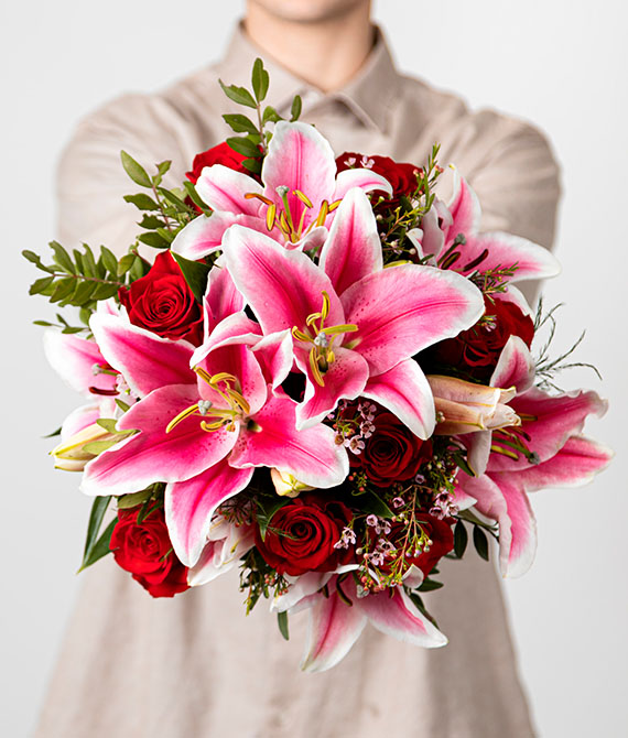 Ramo de rosas rojas y lilium oriental rosa, flor de cera y verdes variados. Envuelto en papel de seda con ilustración de rosas.